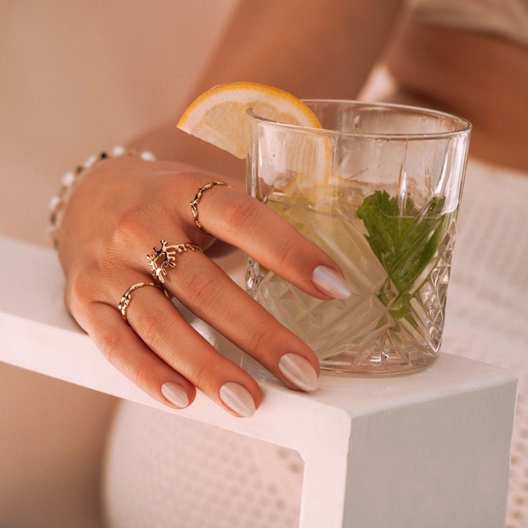 Gros plan main femme portant trois bagues acier inoxydable dorées, une à motif, et verre boisson citron/menthe.