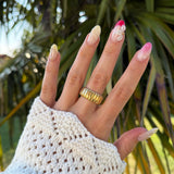 Bague côtelée en acier inoxydable doré portée sur une main féminine avec des ongles décorés et un pull blanc.