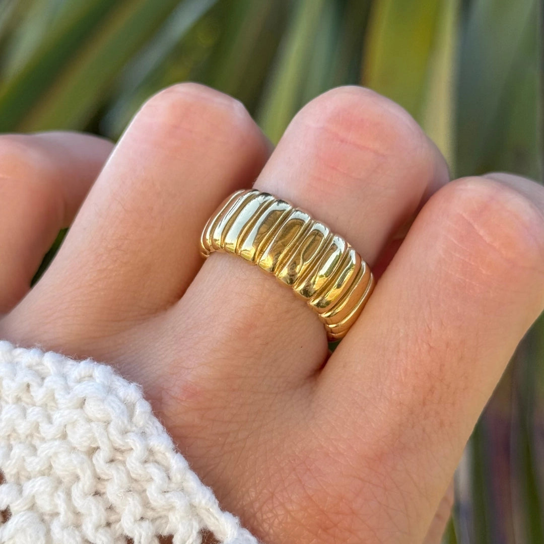 Bague acier inoxydable doré, large design nervuré style croissant, portée sur doigt avec pull blanc.
