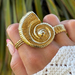 Bague en acier inoxydable doré, grand motif spirale de coquillage nautilus, portée sur une main féminine.