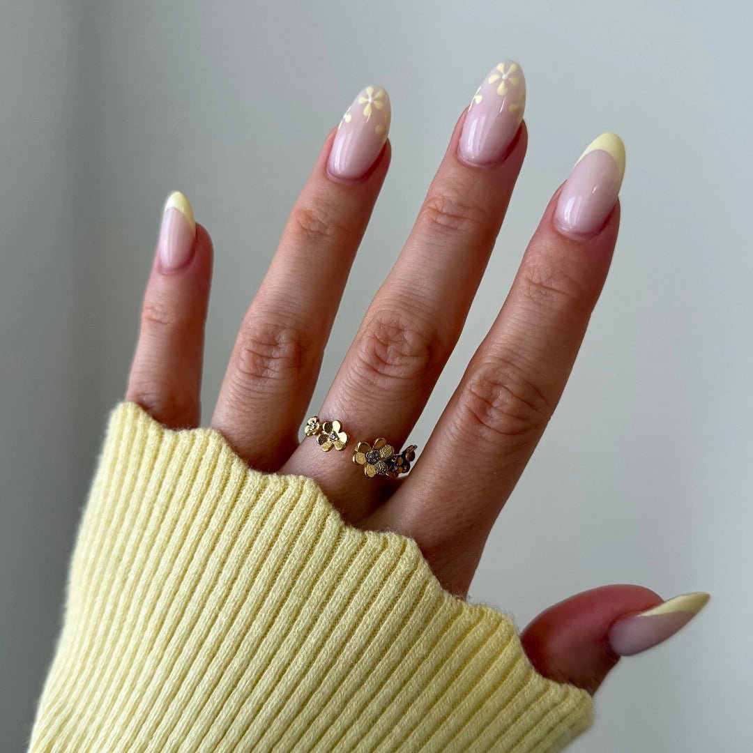 Bague en acier inoxydable doré composée de petites fleurs, portée sur une main au pull jaune et aux ongles fleuris.