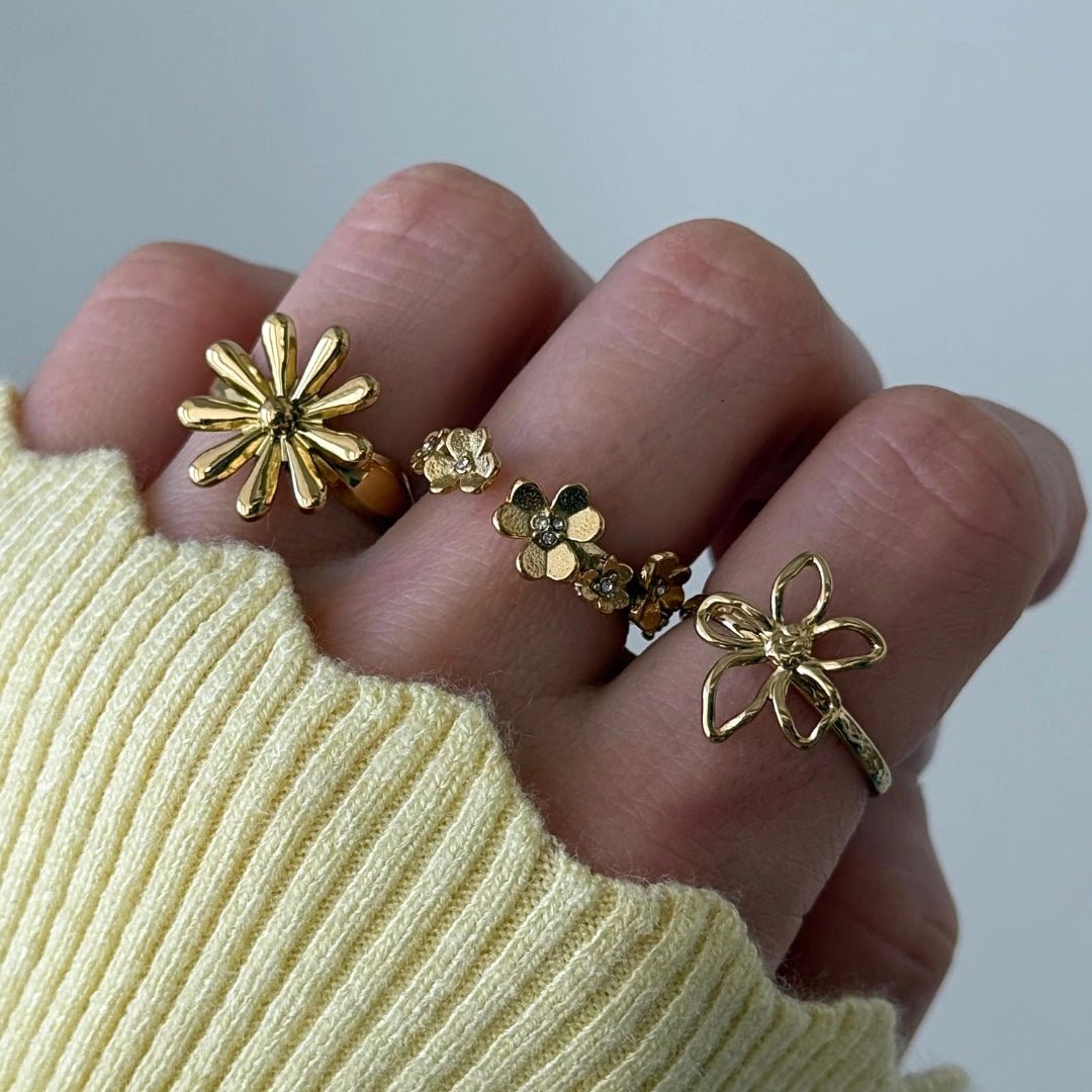Main portant 4 bagues en acier inoxydable doré, motif floral varié: marguerite, petites fleurs avec strass, et fleur ajourée.