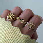 Main portant 4 bagues en acier inoxydable doré, motif floral varié: marguerite, petites fleurs avec strass, et fleur ajourée.