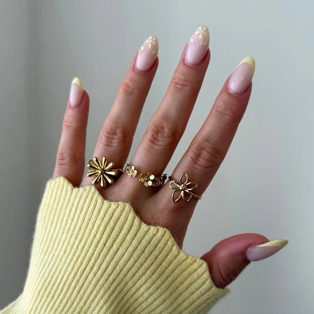 Bagues fleurs en acier inoxydable doré portées sur une main, avec ongles motifs marguerites et pull jaune.