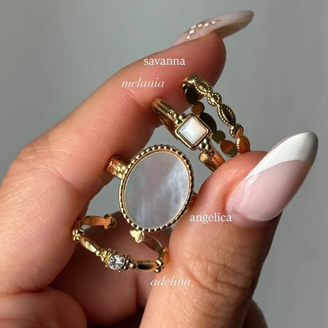 Main tenant une bague ovale en acier inoxydable doré, avec une pierre de nacre texturée. Trois autres bagues dorées sont