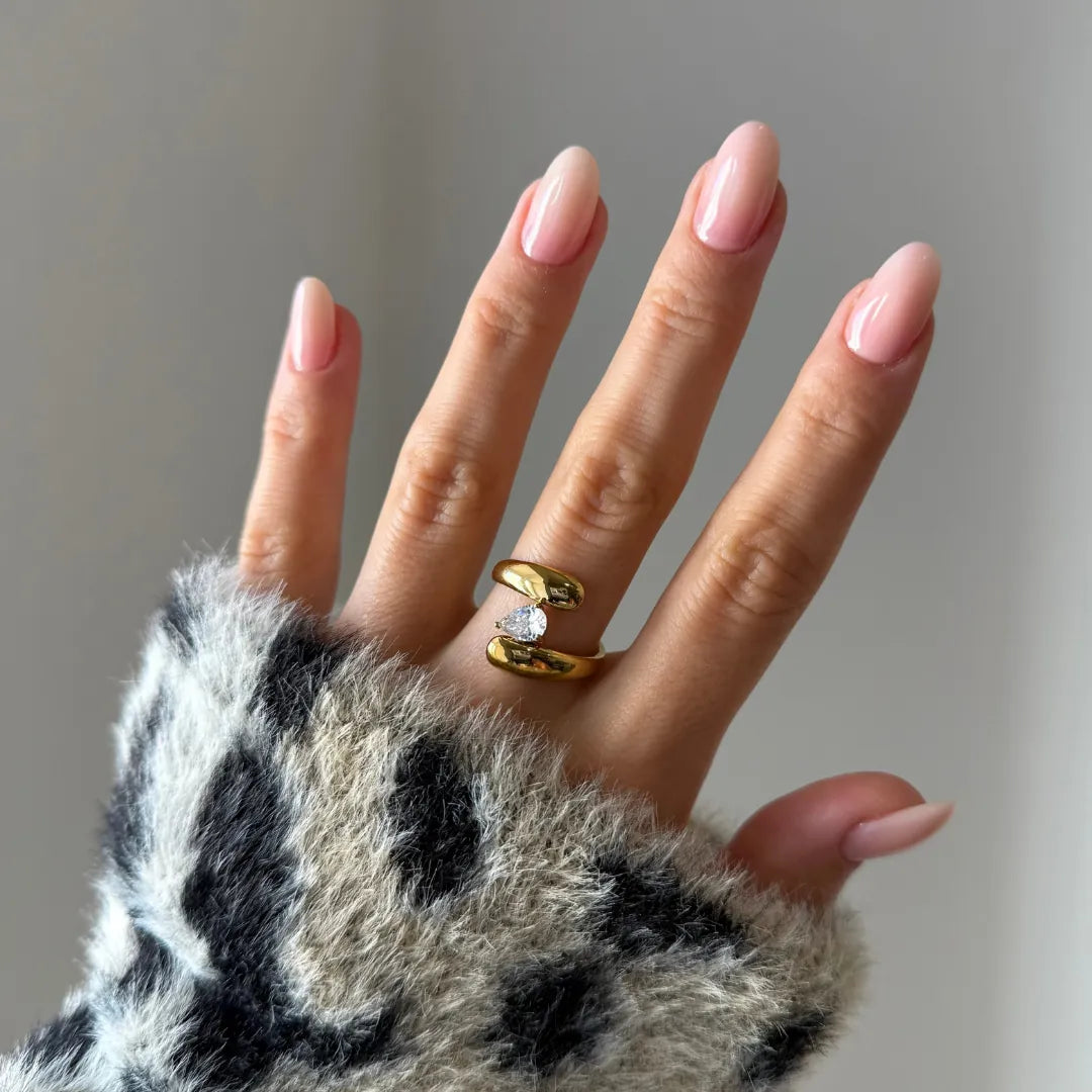 Bague ouverte en acier inoxydable doré, sertie d'un zirconium taille poire, portée sur un doigt à manucure rose, pull doux en