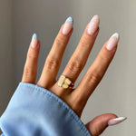 Main avec une bague dorée, sertie de trois pierres claires aux formes variées. Ongles longs french manucure bleue à pois