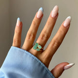 Main portant une bague dorée à trois pierres vertes de formes variées. Ongles vernis aux pointes bleues et points blancs.