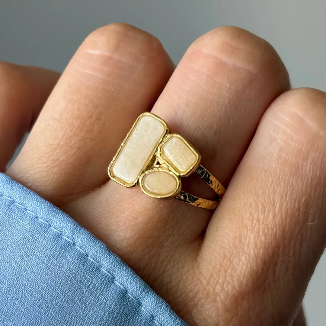 Bague dorée martelée avec trois pierres opaques blanc cassé: une ovale et deux rectangulaires arrondies, portée sur un doigt.