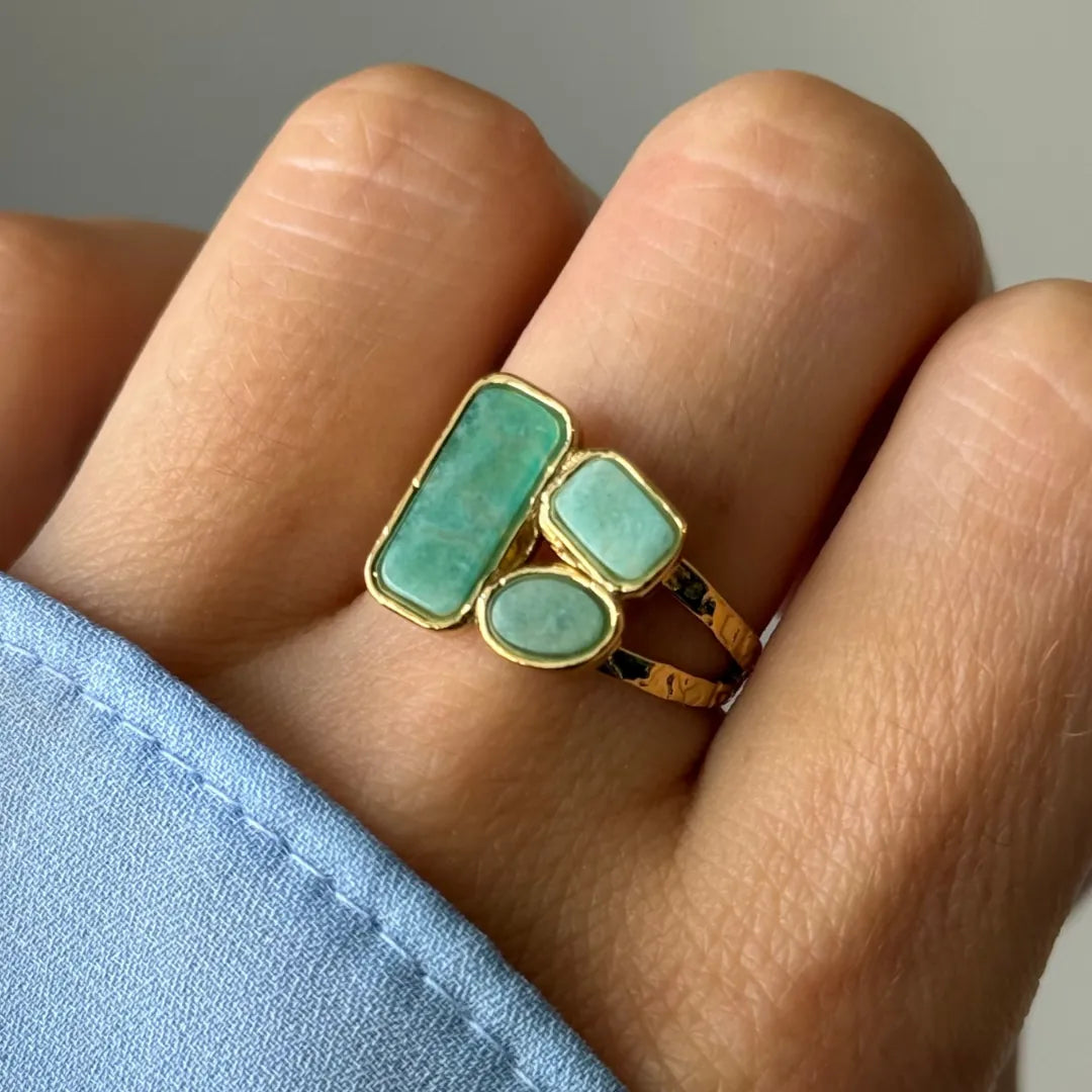 Main portant une bague dorée. Elle a trois cabochons verts aux formes variées (rectangulaire, ovale, arrondie). Double anneau