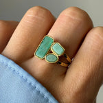 Main portant une bague dorée. Elle a trois cabochons verts aux formes variées (rectangulaire, ovale, arrondie). Double anneau