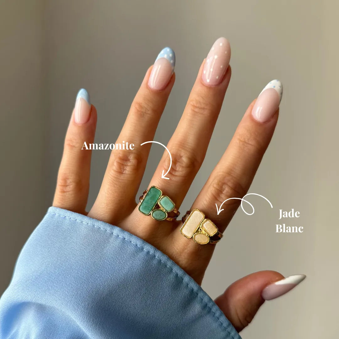 Main portant deux bagues dorées. Une bague amazonite aux pierres vert-bleu, l'autre bague jade blanc aux pierres blanc-crème.