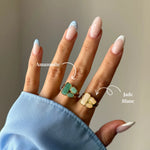 Main portant deux bagues dorées. Une bague amazonite aux pierres vert-bleu, l'autre bague jade blanc aux pierres blanc-crème.