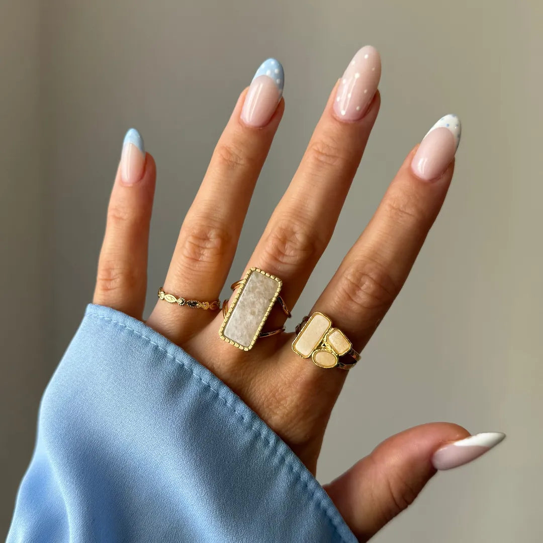 Main de face avec trois bagues dorées: une fine, une rectangle à pierre blanche, une à trois pierres blanches. Manucure