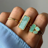 Main portant 3 bagues dorées. Une bague fine ornée, une rectangulaire à pierre vert d'eau, une à 3 pierres pâles et anneau