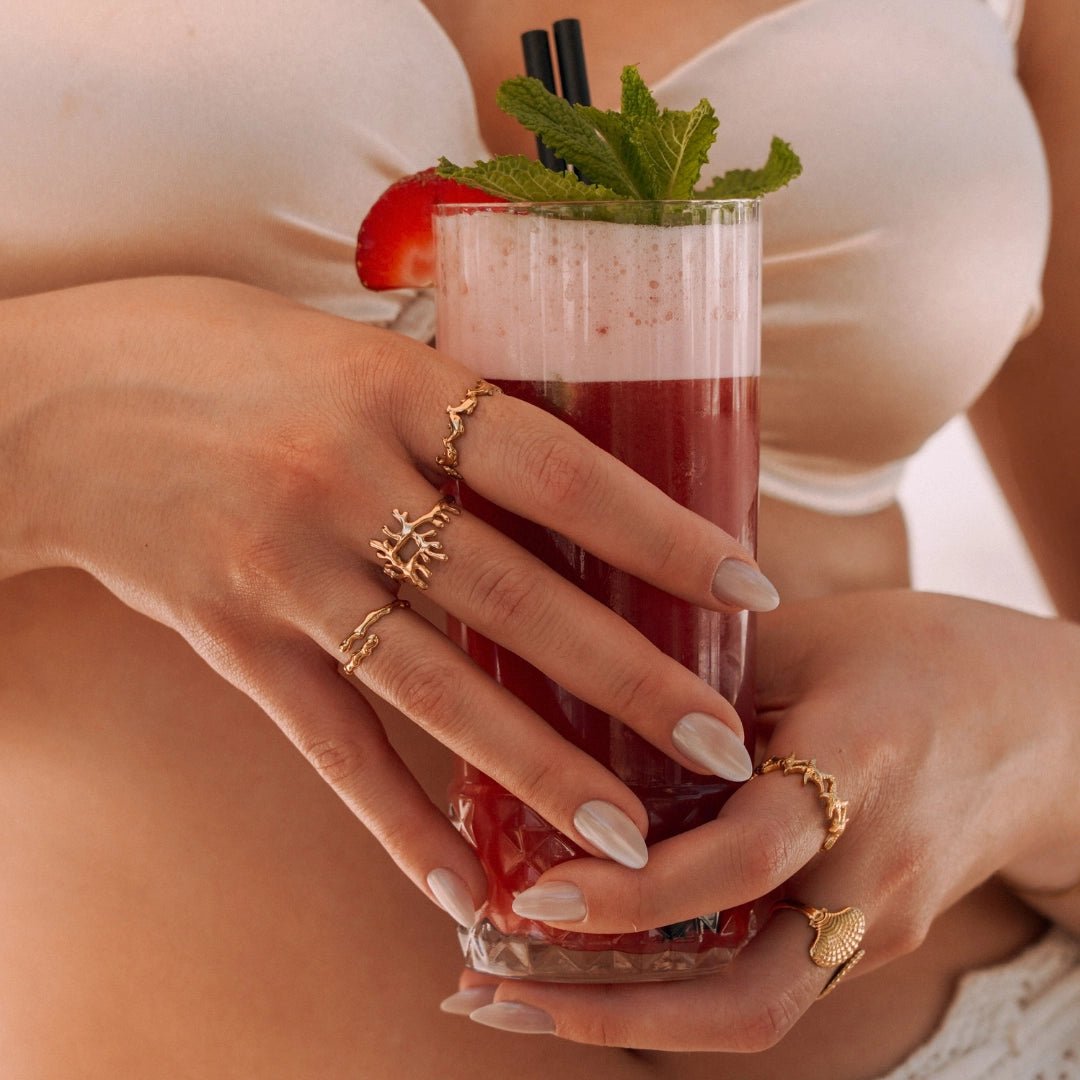 Mains d'une femme portant des bagues en acier inoxydable doré: motif corail, étoile de mer et coquillage, tenant un cocktail.