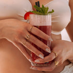 Mains d'une femme portant des bagues en acier inoxydable doré: motif corail, étoile de mer et coquillage, tenant un cocktail.