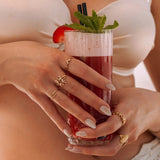 Mains d'une femme portant des bagues en acier inoxydable doré: motif corail, étoile de mer et coquillage, tenant un cocktail.