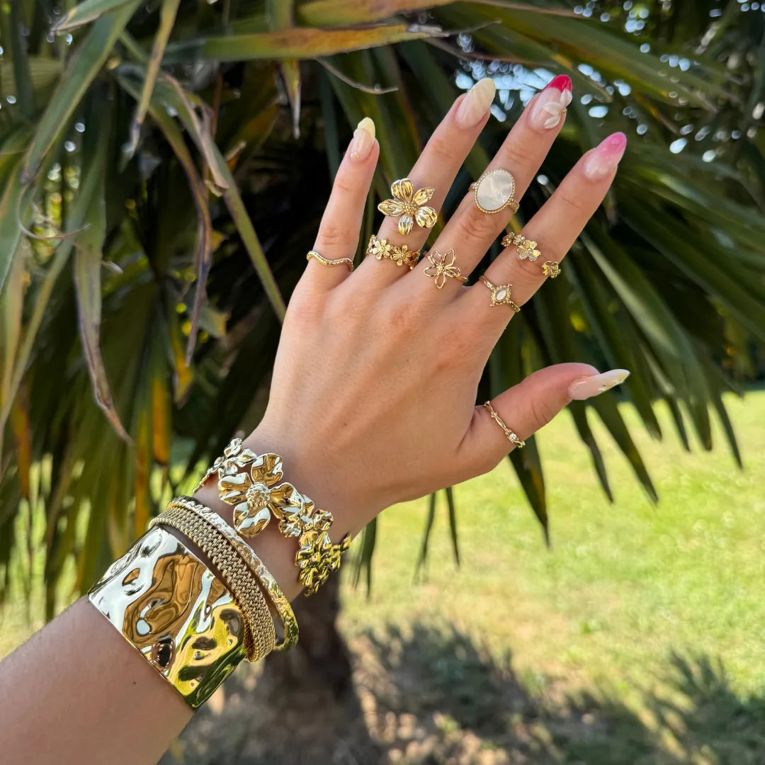 Main de femme avec bagues et bracelets en acier inoxydable doré; une bague fleur de cerisier et une avec cabochon blanc.