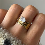 Bague chevalière en acier inoxydable doré martelée organique, avec un zirconium rond, portée sur un doigt.