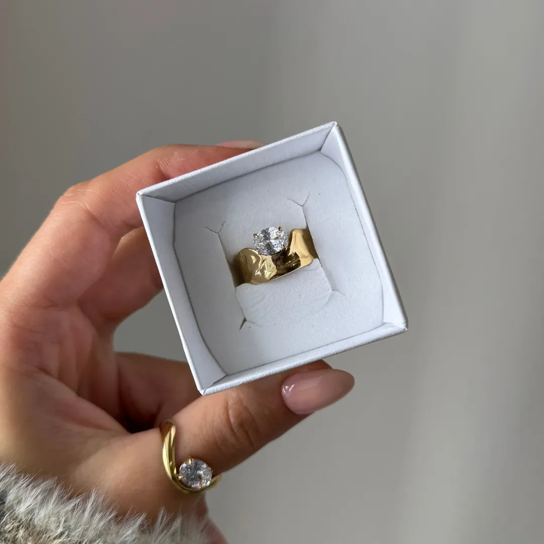 Main tenant une boîte blanche avec bague chevalière en acier inoxydable doré organique martelée et zirconium. Une bague