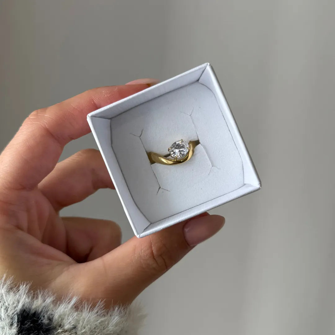 Main tenant une boîte blanche, présentant une bague en acier inoxydable doré ornée d'un Zirconium rond.