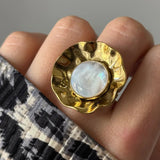 Main portant une bague dorée martelée, design floral, avec un cabochon rond en nacre blanche irisée. Manche à imprimé animal.