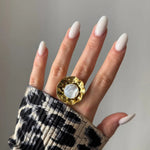 Main de femme aux ongles longs blancs laiteux, bague dorée martelée ondulée avec nacre ronde, manche imprimé léopard.