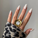 Main de femme aux ongles longs blancs laiteux, bague dorée martelée ondulée avec nacre ronde, manche imprimé léopard.