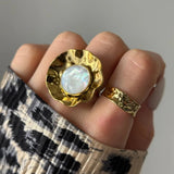 Main avec bague dorée texturée ondulée à nacre irisée centrale, plus une bague dorée martelée. Manchette imprimé léopard.