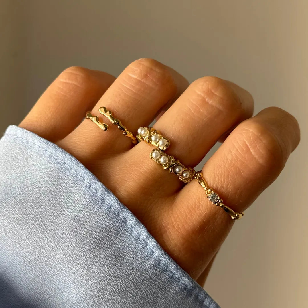 Main sur fond clair, avec une bague ouverte en acier inoxydable doré ornée de perles synthétiques et deux bagues dorées.