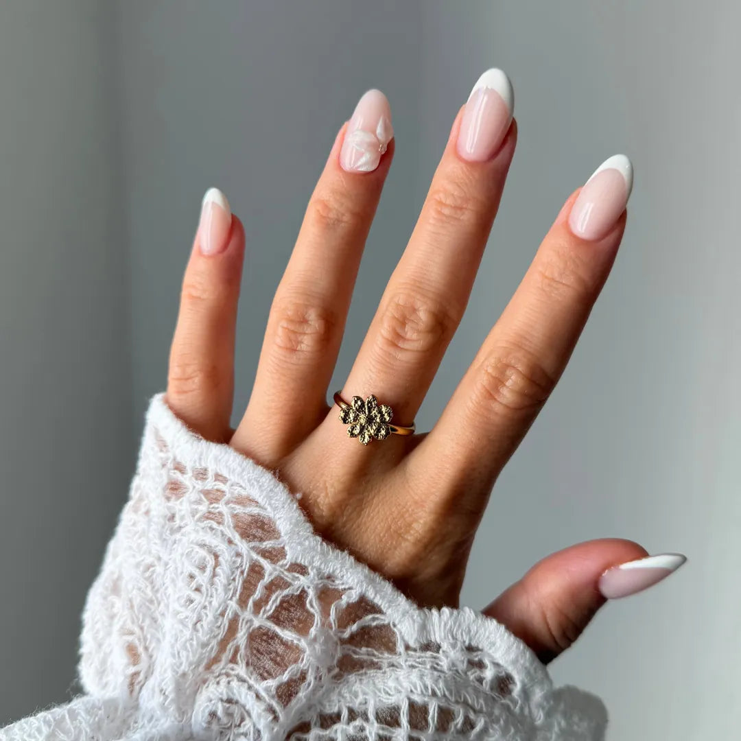Bague fleur texturée en acier inoxydable doré, portée au doigt d'une main à la manche en dentelle blanche.