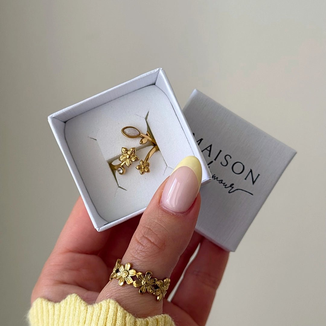 Bague en acier inoxydable doré à motifs fleurs et pierre dans sa boîte cadeau. Une bague fleurie est portée sur le pouce.