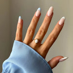 Bague dorée fine avec pierre ovale blanche sur le doigt d'une main aux ongles manucurés bleu et blanc, vêtue d'une chemise