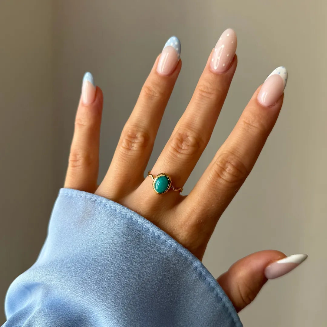 Bague dorée à pierre turquoise ovale portée à la main. Manchette de chemise bleue et ongles en manucure française et pois.