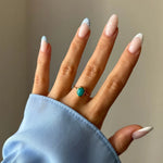 Bague dorée à pierre turquoise ovale portée à la main. Manchette de chemise bleue et ongles en manucure française et pois.