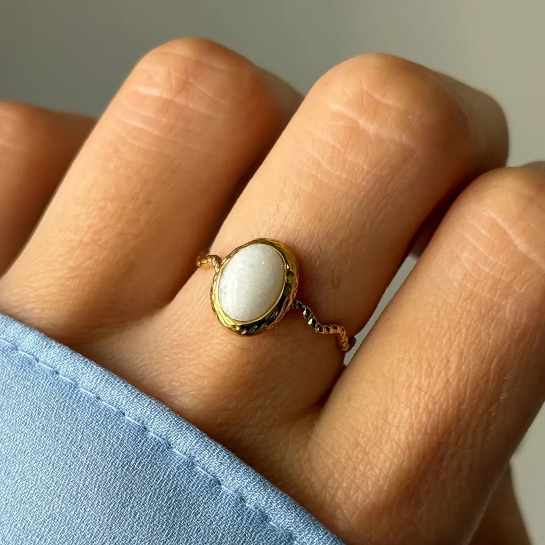 Bague dorée avec une pierre ovale blanche et un anneau torsadé, portée sur un doigt avec une manche bleue.