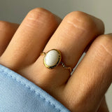 Bague dorée avec une pierre ovale blanche et un anneau torsadé, portée sur un doigt avec une manche bleue.