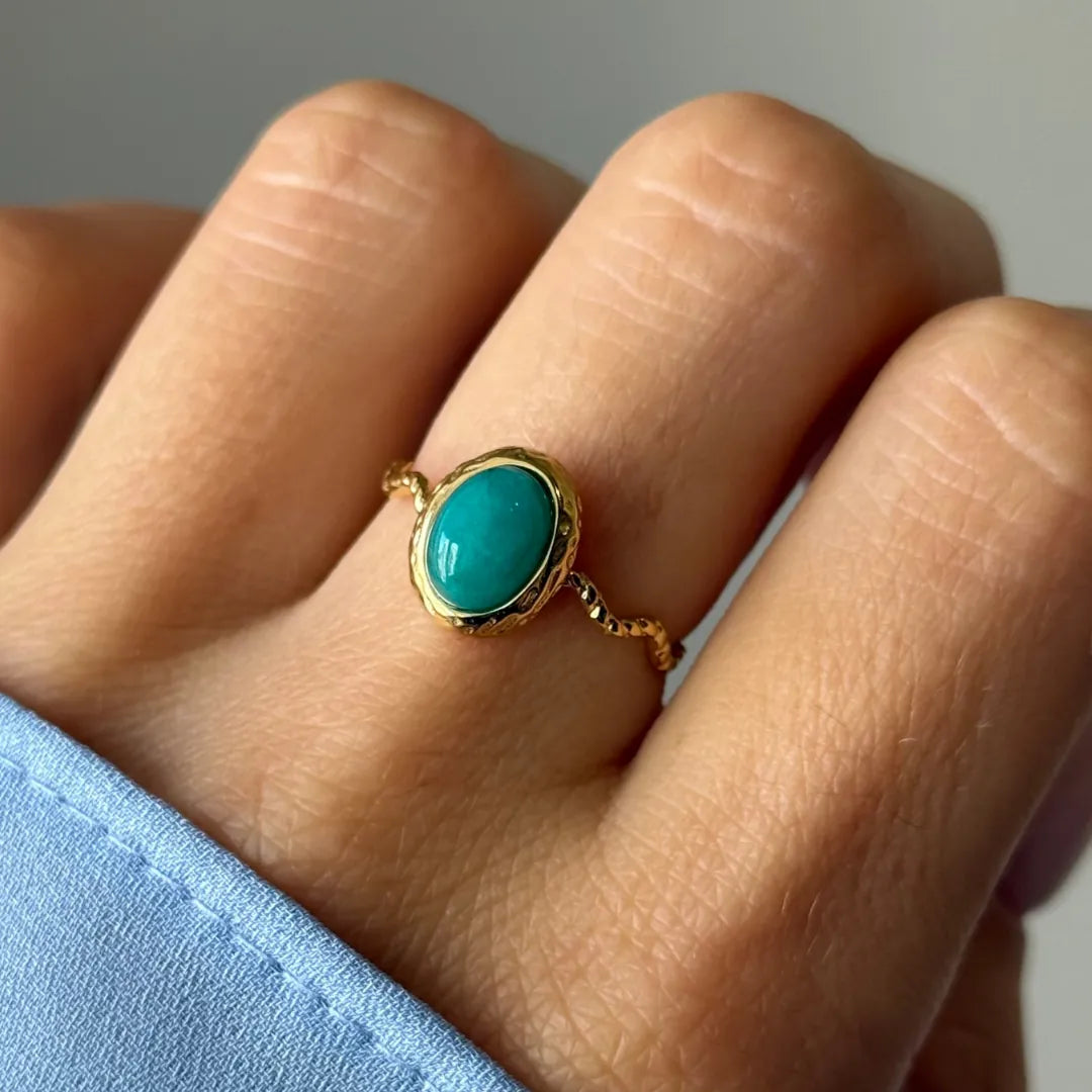 Bague en or portée au doigt. L'anneau est ondulé et orné d'une pierre ovale turquoise avec un serti martelé.