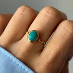 Bague en or portée au doigt. L'anneau est ondulé et orné d'une pierre ovale turquoise avec un serti martelé.
