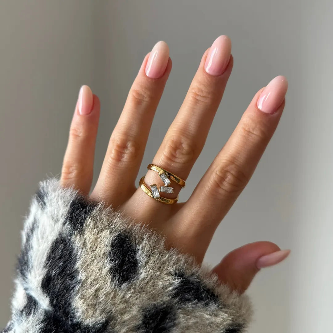 Bague double anneau en acier inoxydable doré à trois pierres baguette en Zirconium, portée sur une main aux ongles roses.
