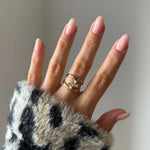 Bague double anneau en acier inoxydable doré à trois pierres baguette en Zirconium, portée sur une main aux ongles roses.