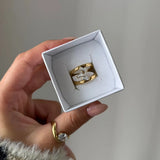 Main tenant un écrin blanc, présentant une bague double anneau en acier inoxydable doré avec trois zirconiums baguette.