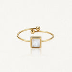 Bague dorée ouverte avec un motif central carré en nacre blanche. Une extrémité est ornée de deux billes dorées, l'autre