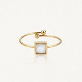 Bague dorée ouverte avec un motif central carré en nacre blanche. Une extrémité est ornée de deux billes dorées, l'autre