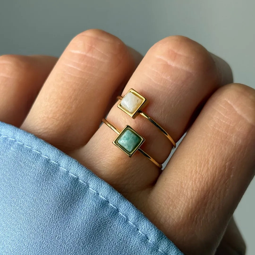 Deux bagues fines dorées avec pierres carrées, une blanche et une verte, portées sur un doigt par une main à manche bleue.