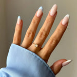 Main portant une bague dorée fine avec une pierre carrée claire. Ongles longs, manucure française bleu ciel et blanche à