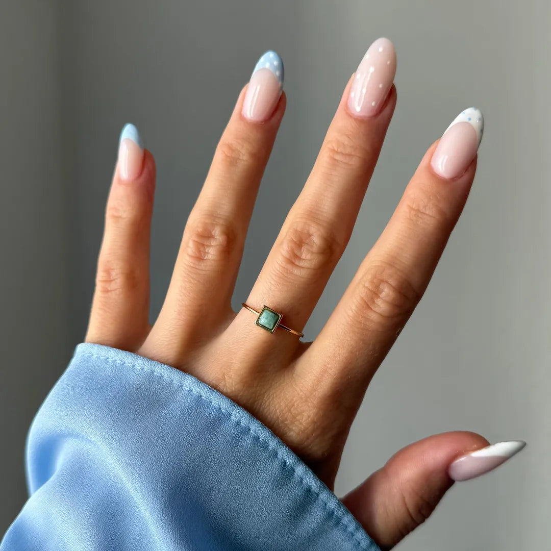 Main portant une fine bague dorée avec une pierre carrée bleu-vert. Ongles vernis nude et bleu ciel à pois blancs.