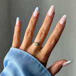 Main portant une fine bague dorée avec une pierre carrée bleu-vert. Ongles vernis nude et bleu ciel à pois blancs.