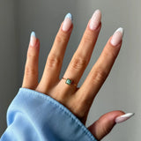 Main portant une fine bague dorée avec une pierre carrée bleu-vert. Ongles vernis nude et bleu ciel à pois blancs.